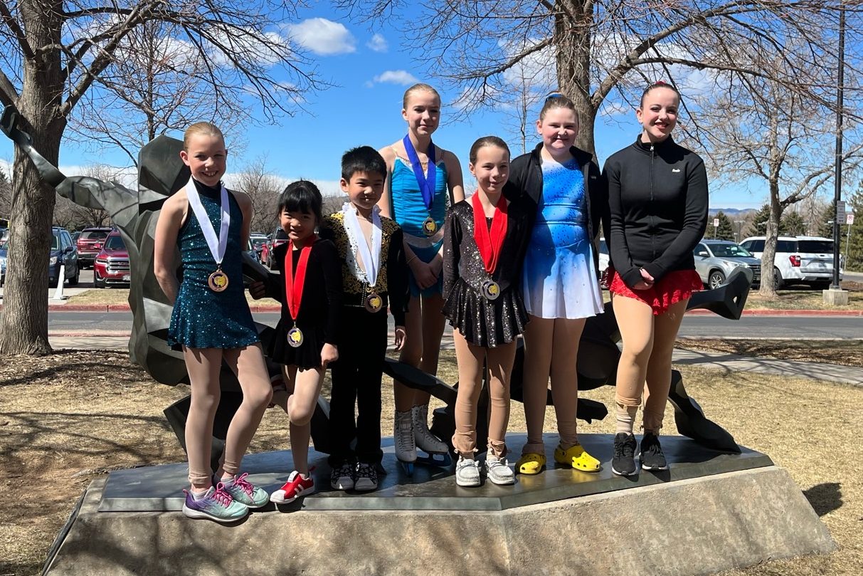 Group of young skaters posing together in front of a statue, wearing medals and skating costumes.