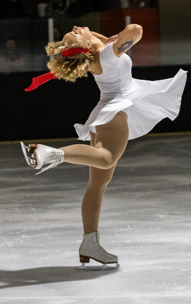 A figure skater performs an elegant spin on ice, wearing a white dress and a red ribbon in her curly hair, showcasing skill and grace.